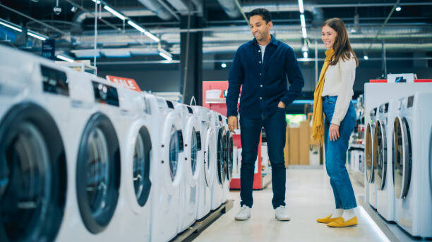 Young Multiethnic Couple Explores Home Electronics Store for Washing Machine Selection. Duo Shopping for Laundry Appliance. Customers Assess Modern Laundry Solutions in Contemporary Retail Shop