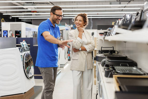 A helpful store assistant guides a customer through various appliances in a modern electronics store. The interaction emphasizes customer service and product knowledge.