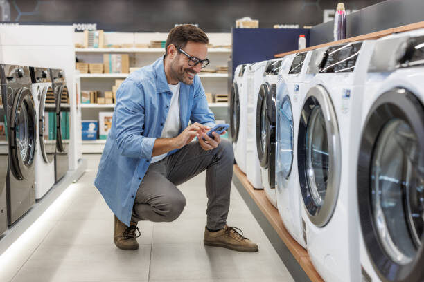 Smiling man crouching as he checks washing machines in a modern appliance store, exploring options and making a purchase decision.